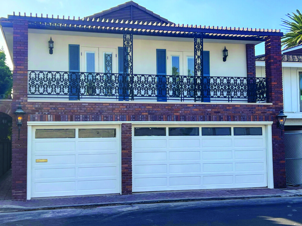 Exterior view of a two-story Newport Beach home featuring white garage doors on the ground level and a second-story balcony with intricate black wrought-iron railings, blue shutters, and red brick accents under a clear blue sky. 