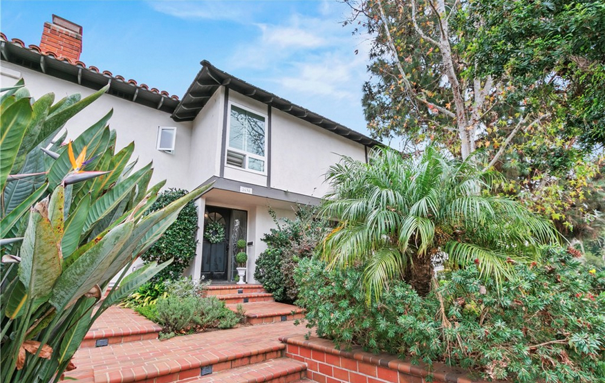 Front exterior of a Spanish-inspired two-story home in Newport Beach featuring a red tile roof, brick entry stairs, and lush Bird of Paradise landscaping.