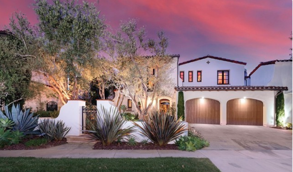 Mediterranean-style Newport Coast home at dusk featuring white stucco walls, arched entryways, a balcony, mature olive trees, and landscaped garden lighting.