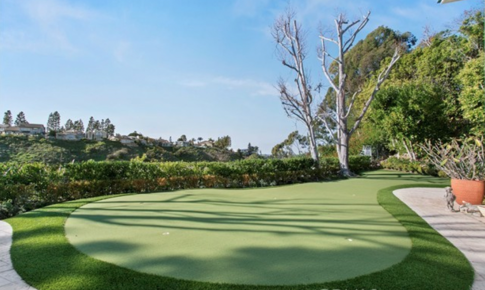 Backyard putting green at a Newport Beach home featuring manicured turf, landscaped hedges, mature trees, and views of surrounding hills.