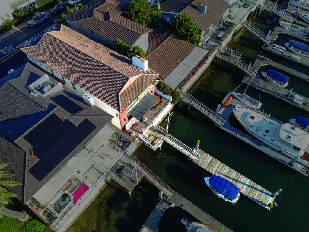 Aerial view of a waterfront home in Newport Beach with a private boat dock extending into the canal, surrounded by neighboring luxury homes and moored yachts.