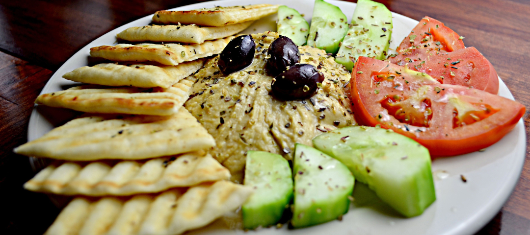 A Mediterranean platter featuring bowls of creamy hummus with olive oil, dark olives, fresh pita bread, sliced cucumbers, and grape tomatoes, overlayed with promotional text for June 2026 Orange County community events.