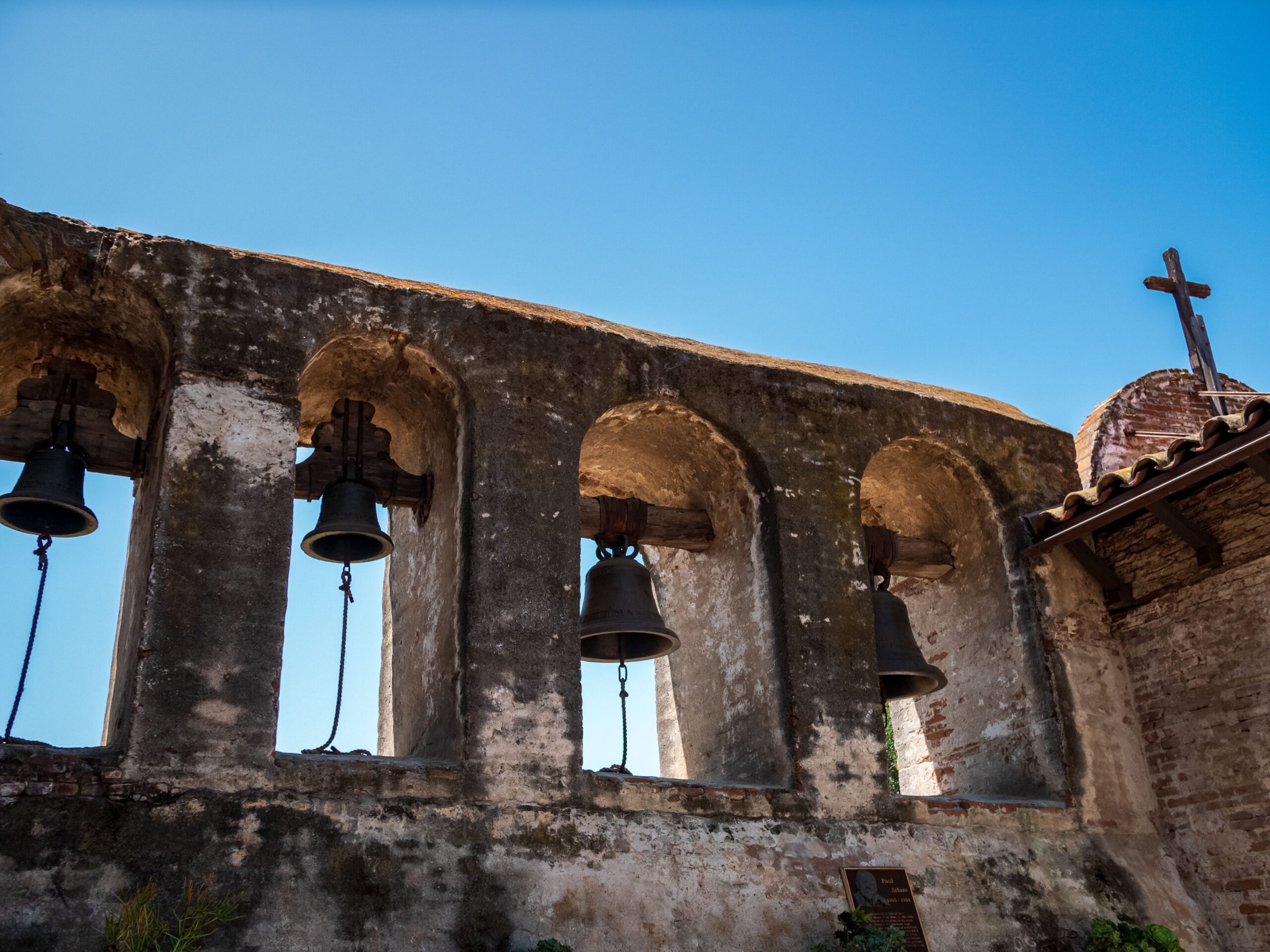 Historic Mission San Juan Capistrano bell wall with arched openings, weathered stone, hanging bells, and blue sky.