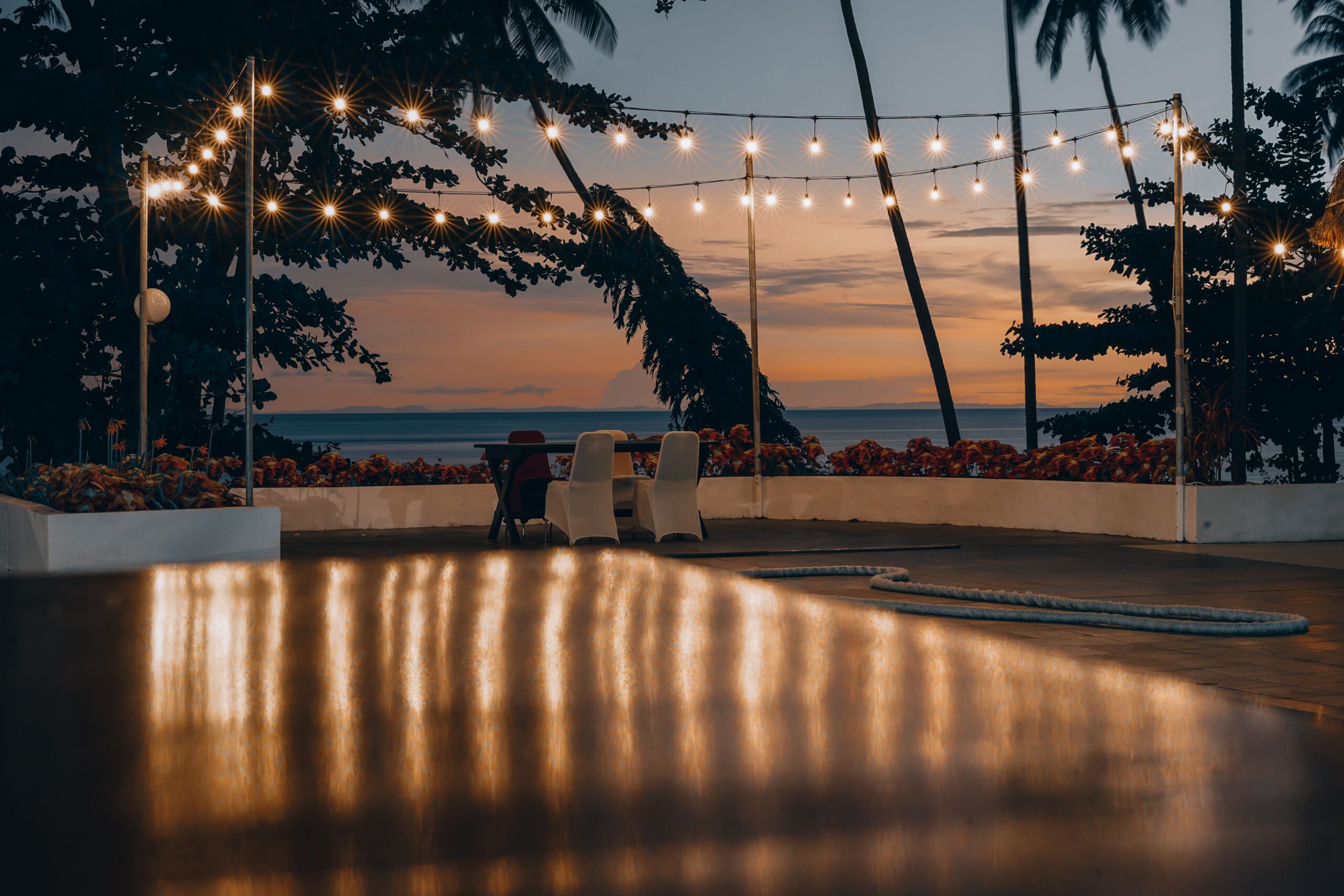 Oceanfront patio at sunset with glowing string lights, palm trees, and views of the Pacific Ocean.