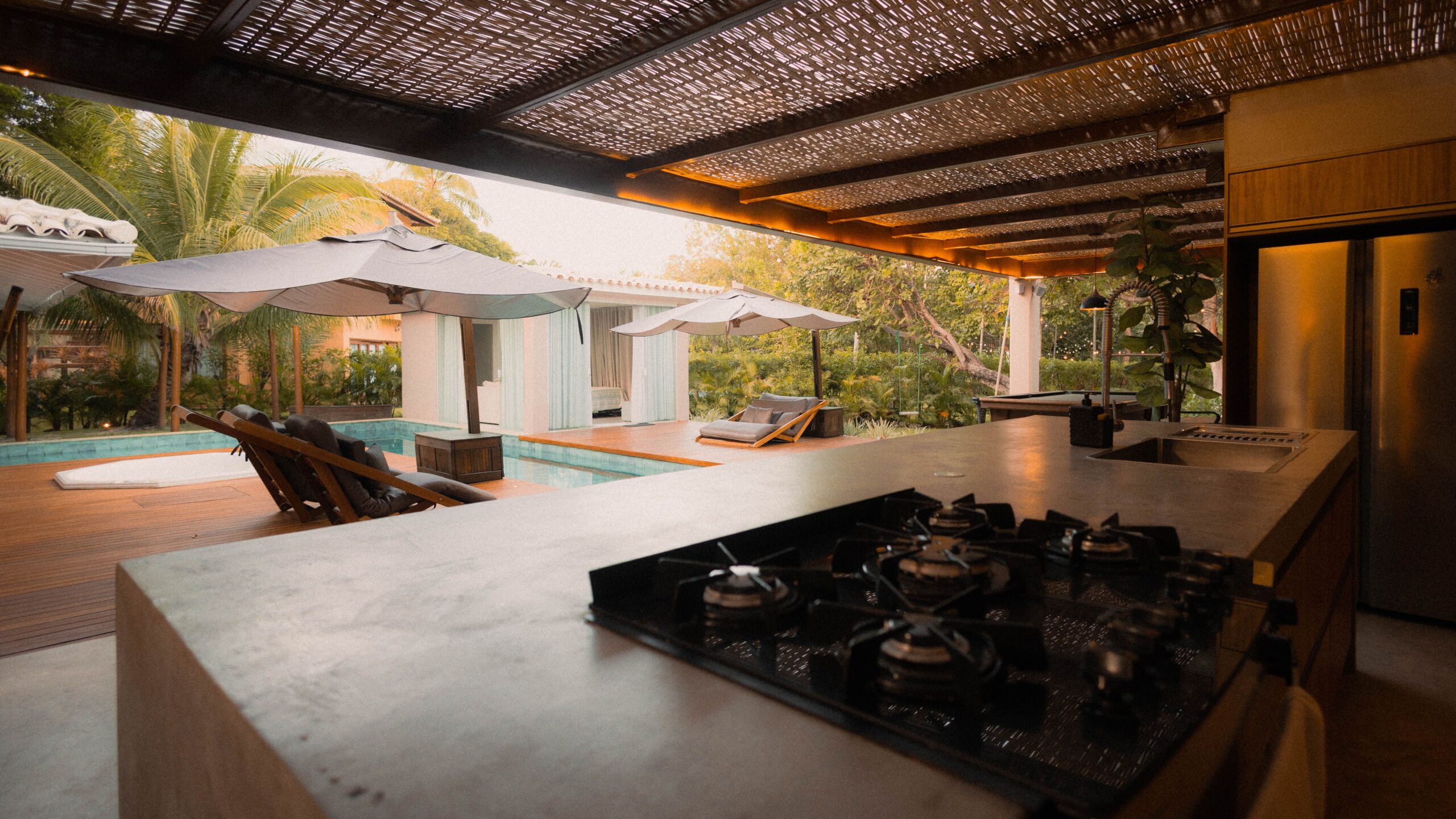 Luxury indoor-outdoor kitchen overlooking a pool, lounge seating, umbrellas, and tropical landscaping.
