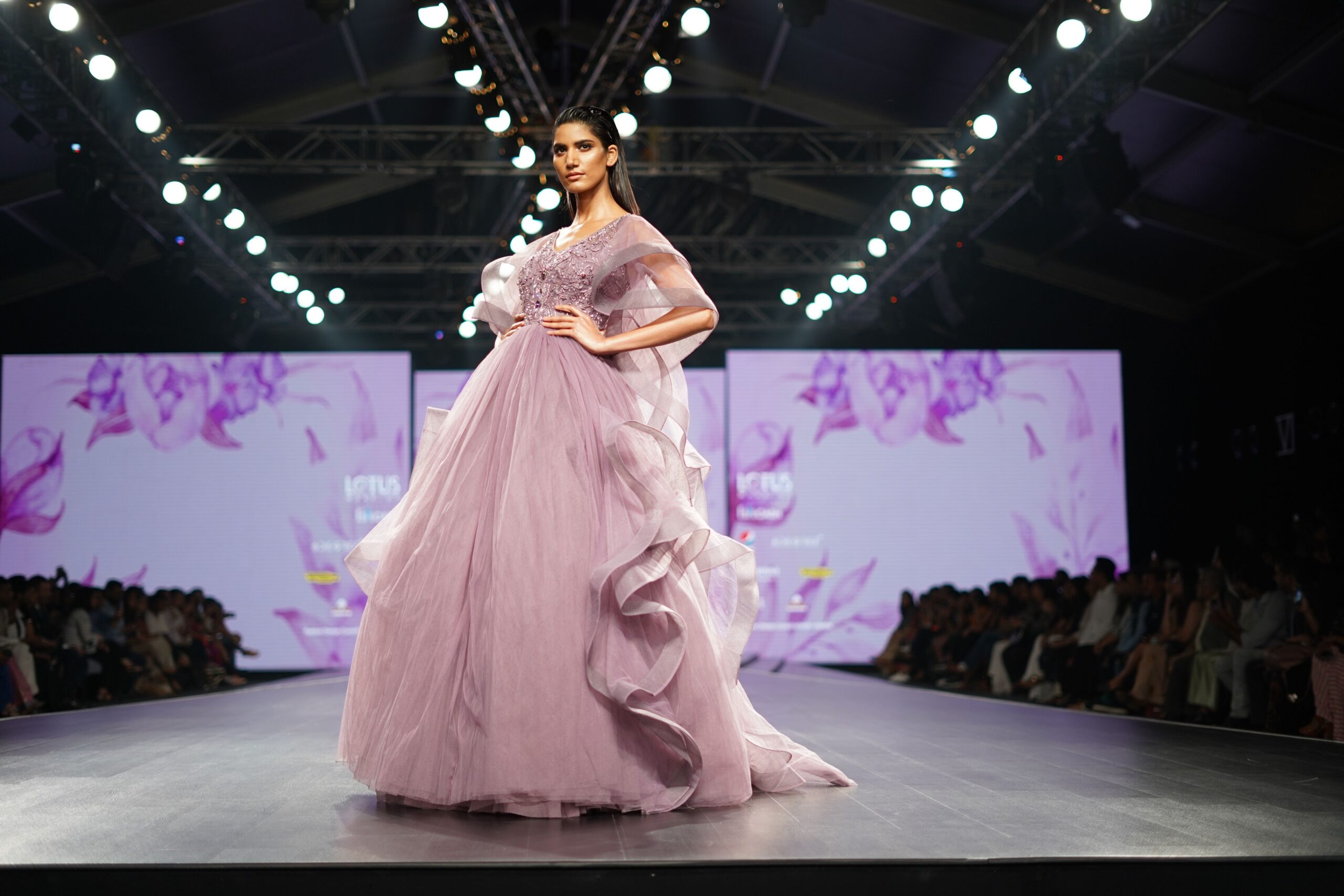 Model in a pink couture gown walking a fashion runway under bright stage lights