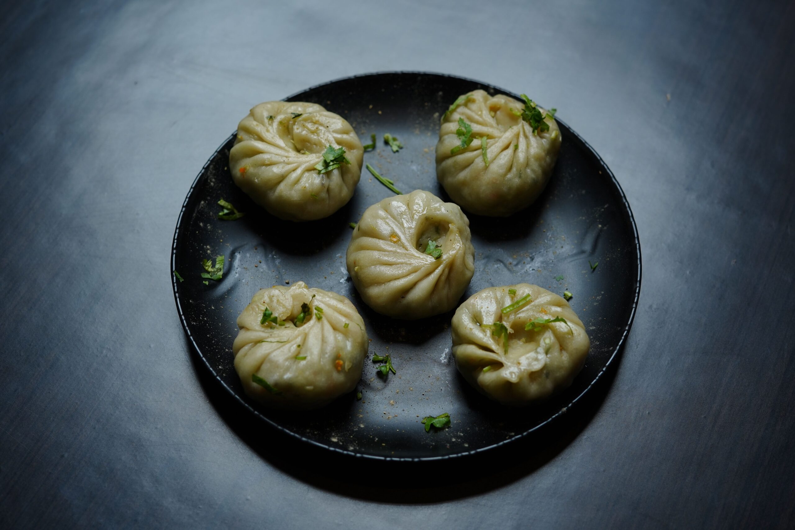 Five steamed dumplings garnished with fresh herbs on a black plate.