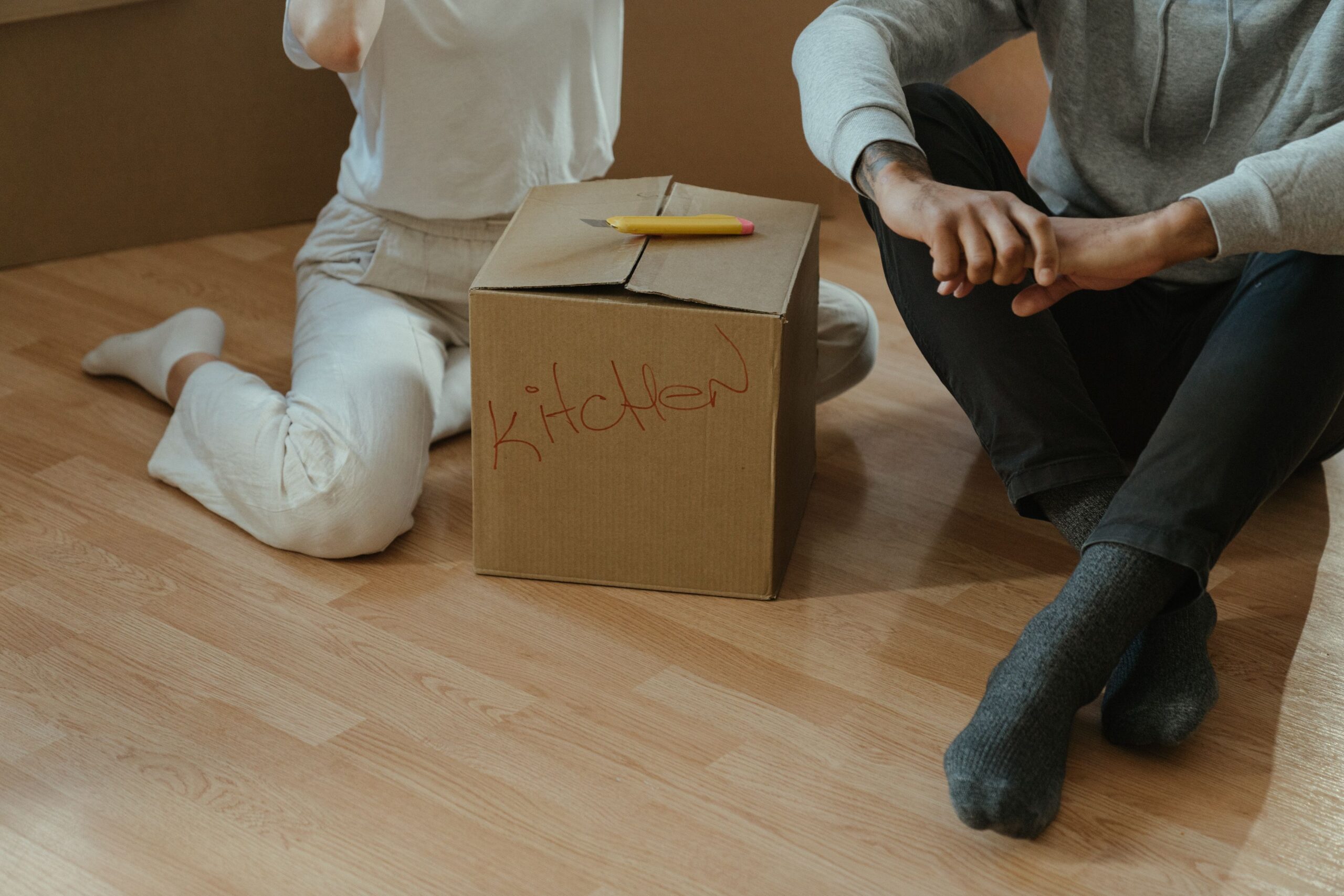 Couple sitting on the floor beside a labeled moving box, representing a home move and changing housing needs.